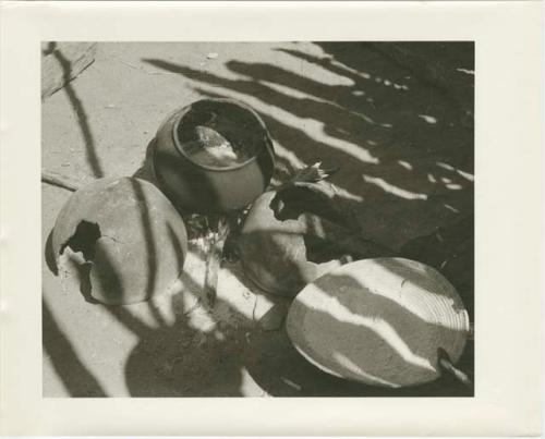 Two broken pots, one whole pot, and a basket holding grain meal; a woman's hand is holding a pestle and the basket (print is a cropped image)