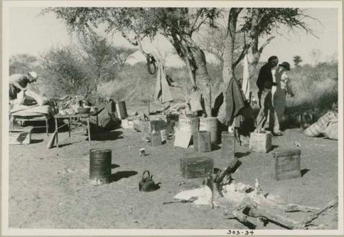 Cooking fire in the expedition camp, with Claude McIntyre with his belongings, Charles Koch and Laurence Marshall walking in the background