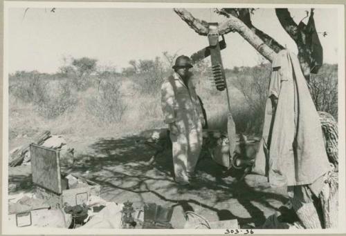 Laurence Marshall standing in the expedition camp next to a cartridge belt hanging from a tree