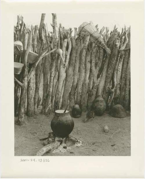 Pot on a fire in a kraal, with baskets hanging from a fence in the background (print is a cropped image)