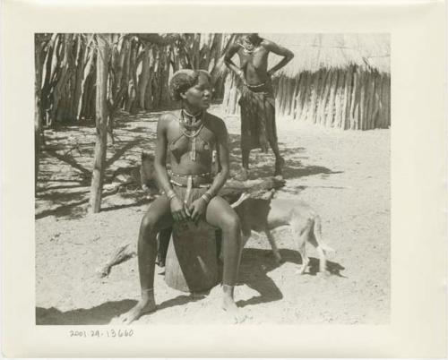 Girl sitting on an upturned mortar, a dog behind her; another girl and a hut in the background (print is a cropped image)