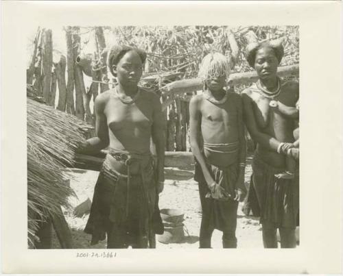 Three girls standing by a hut; one of the girls wears a beaded headdress (print is a cropped image)