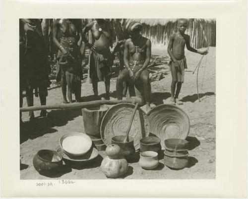 Pots, baskets, and gourds in the foreground; people in the background including a seated girl and a boy with a bow and arrow (print is a cropped image)