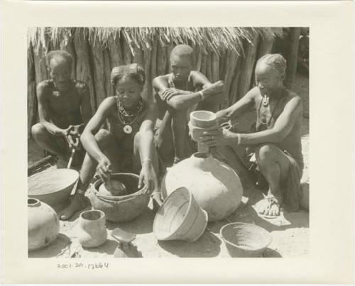 Two women and two men, sitting among gourds and grain baskets; one woman holds a funnel over the mouth of a gourd while the other woman scoops meal with a gourd ladle (print is a cropped image)