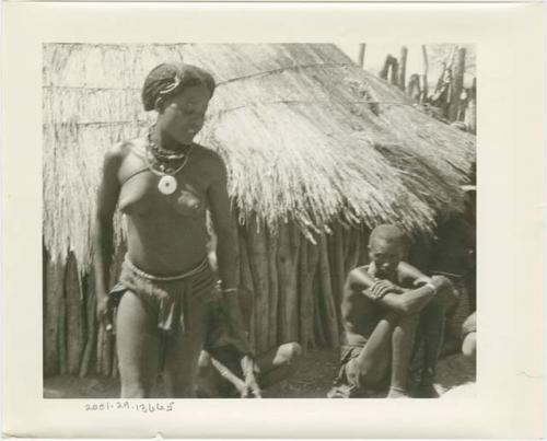 Girl standing beside a hut; other people seated in the background