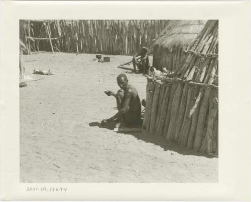 Two men sitting in front of huts in a kraal (print is a cropped image)