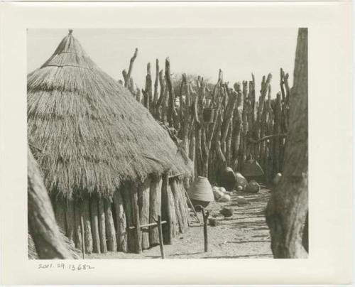 Hut with a door locked by a pole; baskets and pots by fence in the background (print is a cropped image)