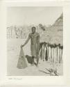 Man standing by a hut and holding a fish basket (print is a cropped image)