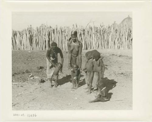 Two women holding children while other children watch; wood pole fence in the background (print is a cropped image)