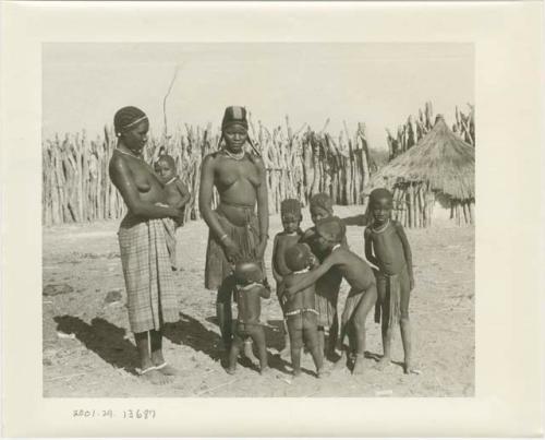 Two women and seven children, one held by a woman; wood pole fence in the background (print is a cropped image)