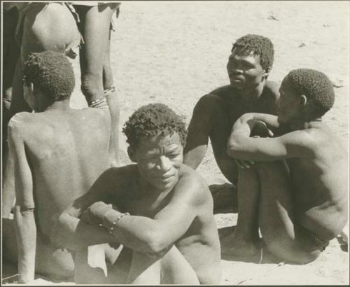 ≠Toma sitting with three other men by a metal drum, waiting to have their plaster cast face molds made (print is a cropped image)