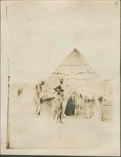 Man and woman standing outside flounced roof hut, child in the entrance, near the Nile River