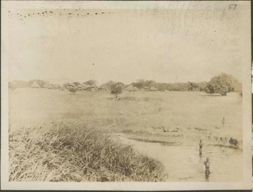 Two people standing in water, with a village in the background surrounded by sparse vegetation and grassland