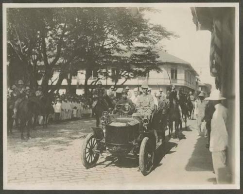 Military procession, one automobile and men on horseback with crowds watching.