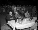 Group of people standing behind Colonel Hoogenhout, Laurence Marshall and John Neser sitting at a table