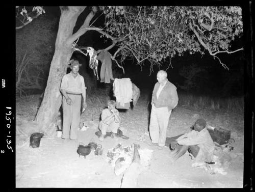 Expedition members sitting and standing in camp, including Edward Hartley, Laurence Marshall and John Marshall, with meat hanging in a tree behind them