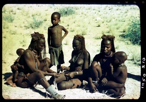 Group of women making a necklace, with children sitting and standing with them