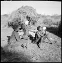 /Gunda (N!ai's husband) steadies a child while the child drinks from an ostrich eggshell, with a woman sitting next to them also drinking from an eggshell