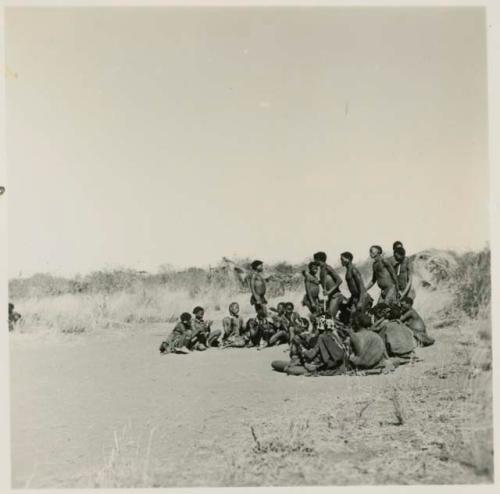 Group of men performing a curing dance, cutting across a circle of women who are clapping and singing