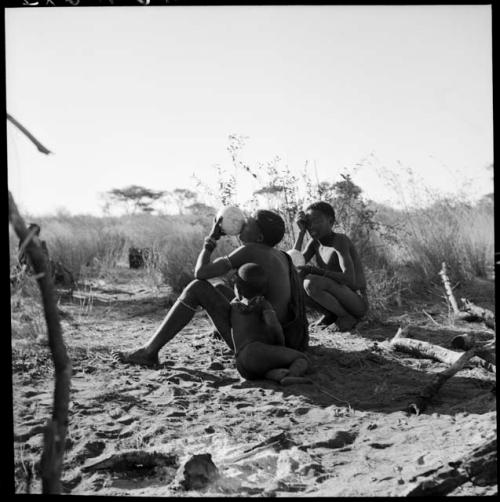 Woman sitting and drinking from an ostrich eggshell, with a child leaning against her, /Gunda sitting behind her