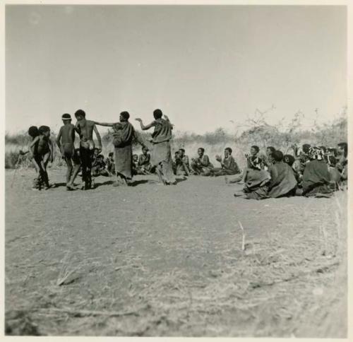 Two women and boys dancing, with a group of women sitting and clapping at the beginning of a dance