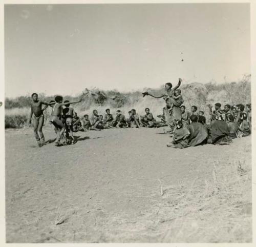 Two women and boys dancing, with a group of women sitting and clapping at the beginning of a dance