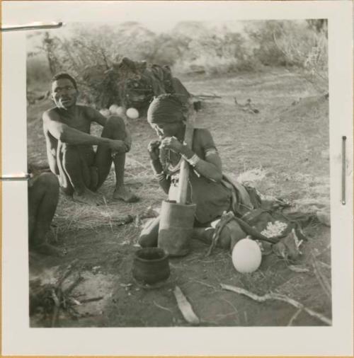 "Gao Medicine" sitting on grass, and Di!ai sitting by the fire and eating something she has pounded in the mortar