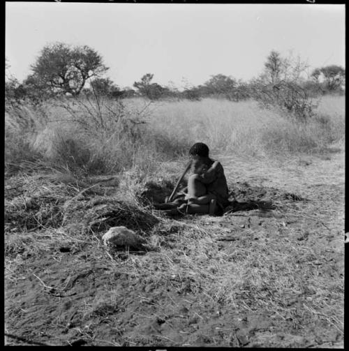 Woman digging with a digging stick, sitting with a child on her leg