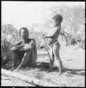 Bo sitting and looking at his daughter Be, who is standing next to him holding a stick