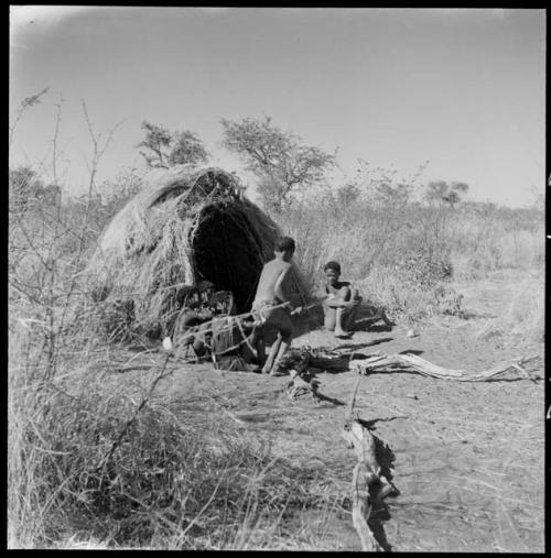 "Little N!ai" and /Gunda (son of Khwan//a and her former husband) sitting by their skerm with their wedding party, including Tsamgao (son of ≠Toma and !U), ≠Gisa (daughter of ≠Gao and Khwo//o-/Gasa), Xama (daughter of "Gao Helmet" and //Kushay), ≠Gao (son of Gau and Be), and a boy playing the //guashi