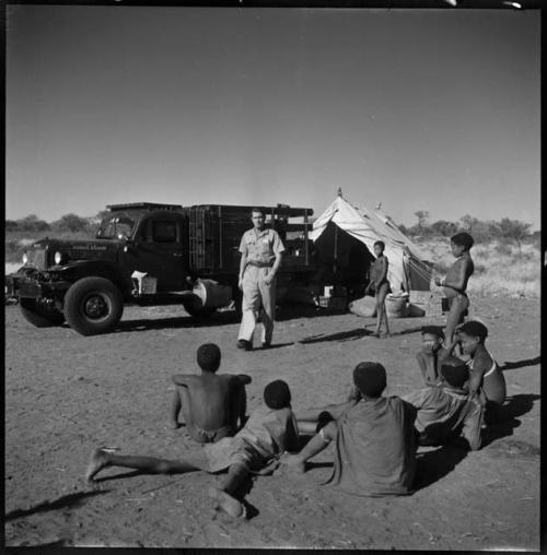 Group of people sitting and standing, with Heiner Kretzchmar walking in front of an expedition truck in the background