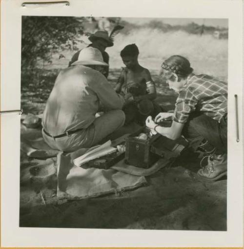 /Naoka holding her daughter /Khoa ("/Qui Hunter's" daughter), sitting with Laurence Marshall and Lorna Marshall, with expedition medicine box visible