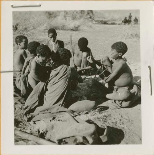 Group of people sitting around a fire, including /Naoka ("Gao Medicine's" first wife) cooking, her son /Gaishay playing a //guashi, "≠Gao Lame," and Gao (son of ≠Gao and Khwo//o-/Gasa) playing a //guashi