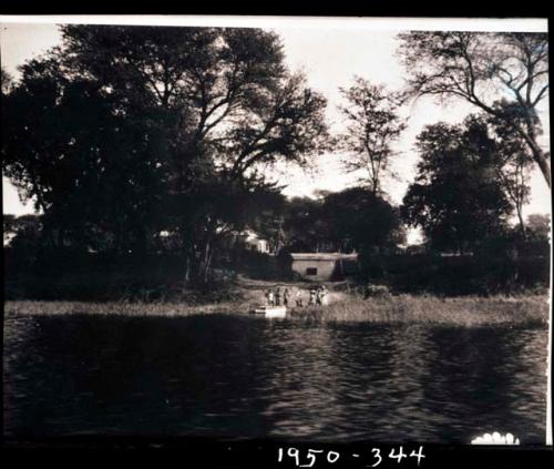 People standing along the shore of the Okavango River, with a building in background