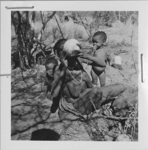 /Naoka (wife of Ti!kay) drinking from an ostrich eggshell, with !Nani behind her, and /Khoa (her daughter) standing beside her