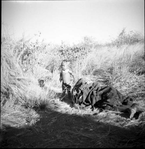 Woman lying on the ground wrapped in a kaross, with Khuan//a (daughter of //Ao and ≠Nisa) standing next to her