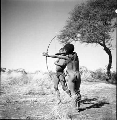 Boy aiming a bow and arrow, with another boy standing next to him