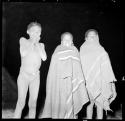 Three boys standing, two wrapped in blankets, illuminated by flashlight at night