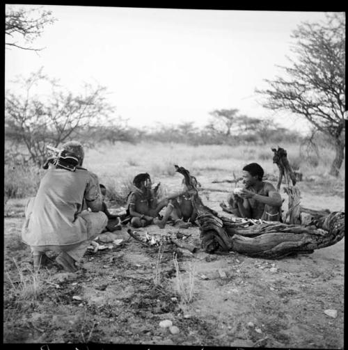 Group of people sitting, with John Marshall crouched next to them, his camera on his back