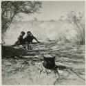 Two people sitting by a fire in the shade, with a pot of mangetti nuts cooking in front of them