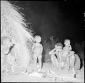 Two women sitting next to a fire in front of a skerm at night, with two children standing next to them