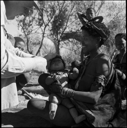 Khuan//a (daughter of ≠Gao and Khwo//o-Gasa of Band 4) sitting, holding her baby, with an expedition member looking at the baby