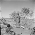 //Kushay sitting, watching !Ungka hanging her bag of possessions in a tree, view from behind