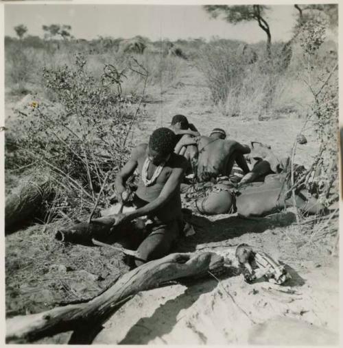 Man making a bag from the whole skin of a wild pig, with a group of men sitting behind him