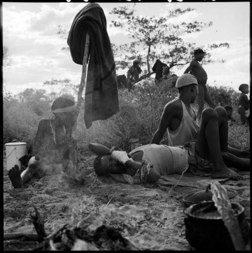 Two boys playing, with a girl sitting next to them watching, pot of food cooking on a fire near them