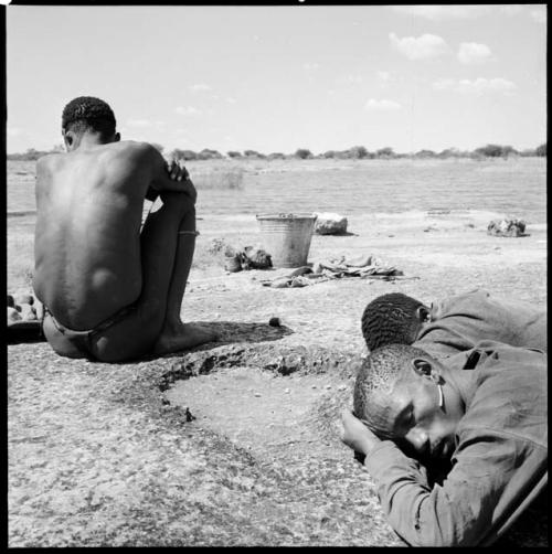 Man sitting at the edge of a pan, view from behind, with two men sleeping next to him, bucket on the ground in front of them