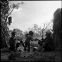 N!ai offering food to two children, sitting with another woman between two skerms