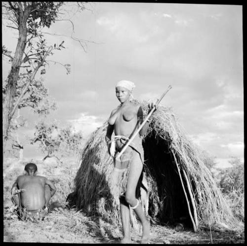 Woman wearing a modest apron, holding a digging stick, standing in front of her skerm, with her husband sitting beside the skerm, facing away from the camera