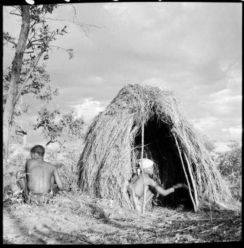 Woman squatting inside her skerm, with her husband sitting beside the skerm, facing away from the camera