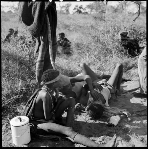 Two boys playing, with a girl sitting next to them watching, enamel pot on the ground next to her, blankets hanging on a post behind them